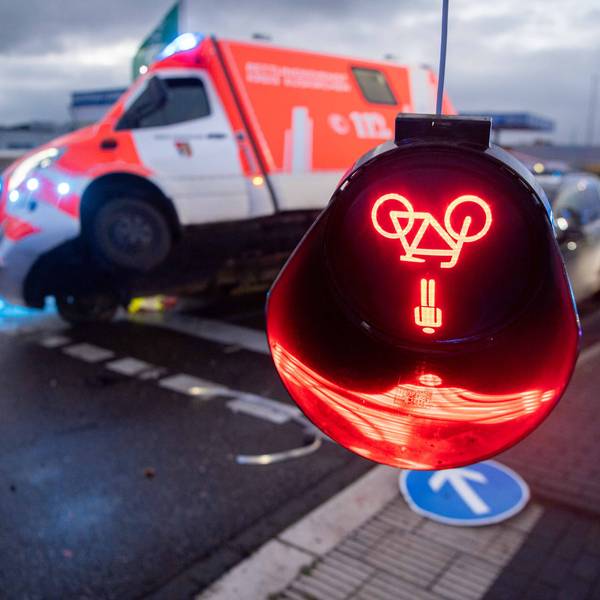 Das Symbolfoto zeigt eine bei einem Unfall beschädigte Ampel, deren rotes Leuchtzeichen auf dem Kopf hängt. Im Hintergrund ist ein beschädigter Rettungswagen zu sehen.