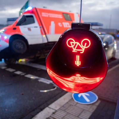 Das Symbolfoto zeigt eine bei einem Unfall beschädigte Ampel, deren rotes Leuchtzeichen auf dem Kopf hängt. Im Hintergrund ist ein beschädigter Rettungswagen zu sehen.