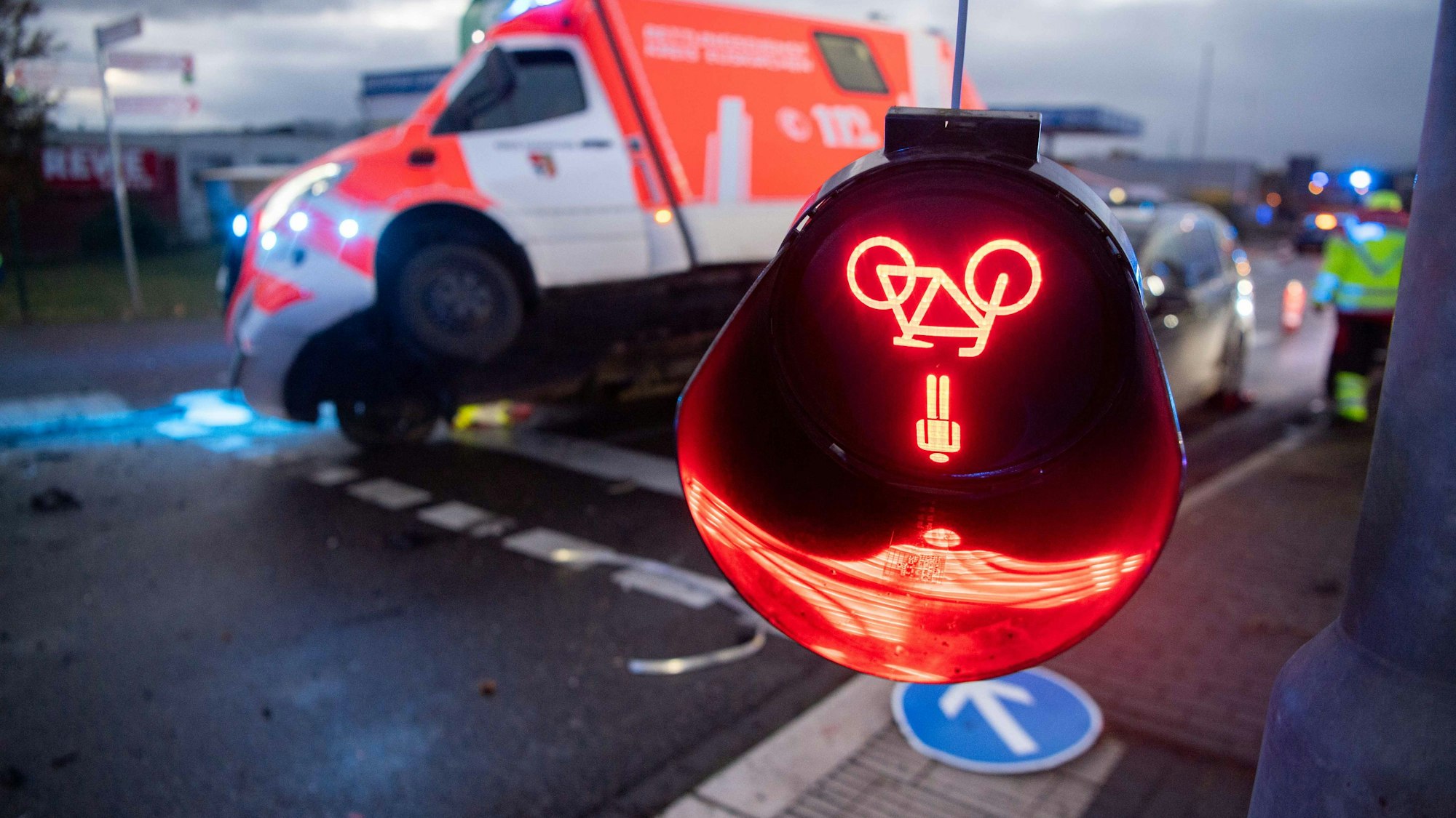Das Symbolfoto zeigt eine bei einem Unfall beschädigte Ampel, deren rotes Leuchtzeichen auf dem Kopf hängt. Im Hintergrund ist ein beschädigter Rettungswagen zu sehen.
