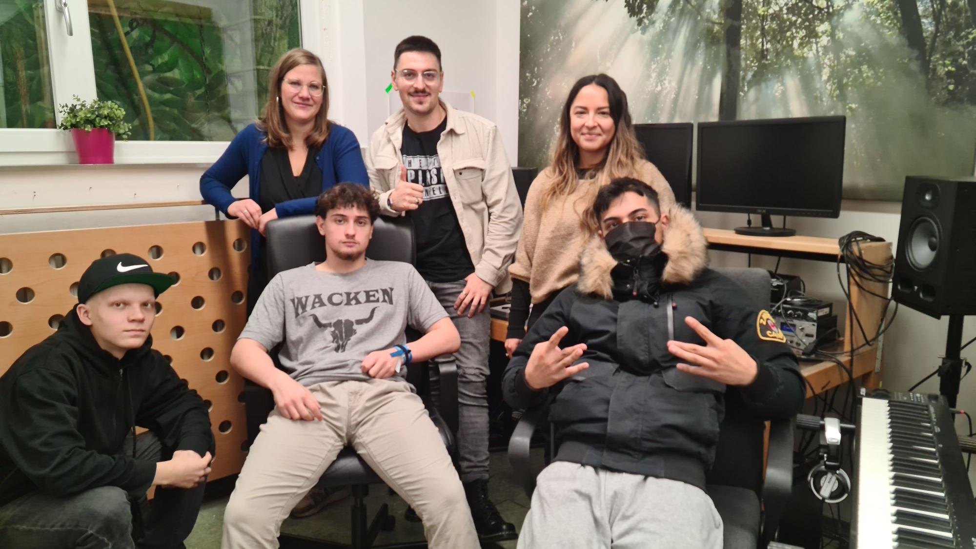 Vier junge Männer und zwei Frauen stehen und sitzen in einem Tonstudio.