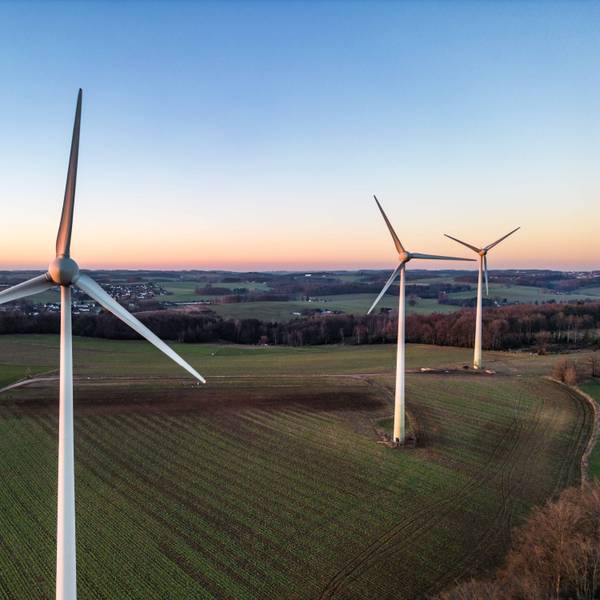 Drei große Windräder stehen auf einem Feld, schmale Straßen und ein Waldgebiet rahmen das Feld ein, im Hintergrund liegt eine kleine Siedlung.