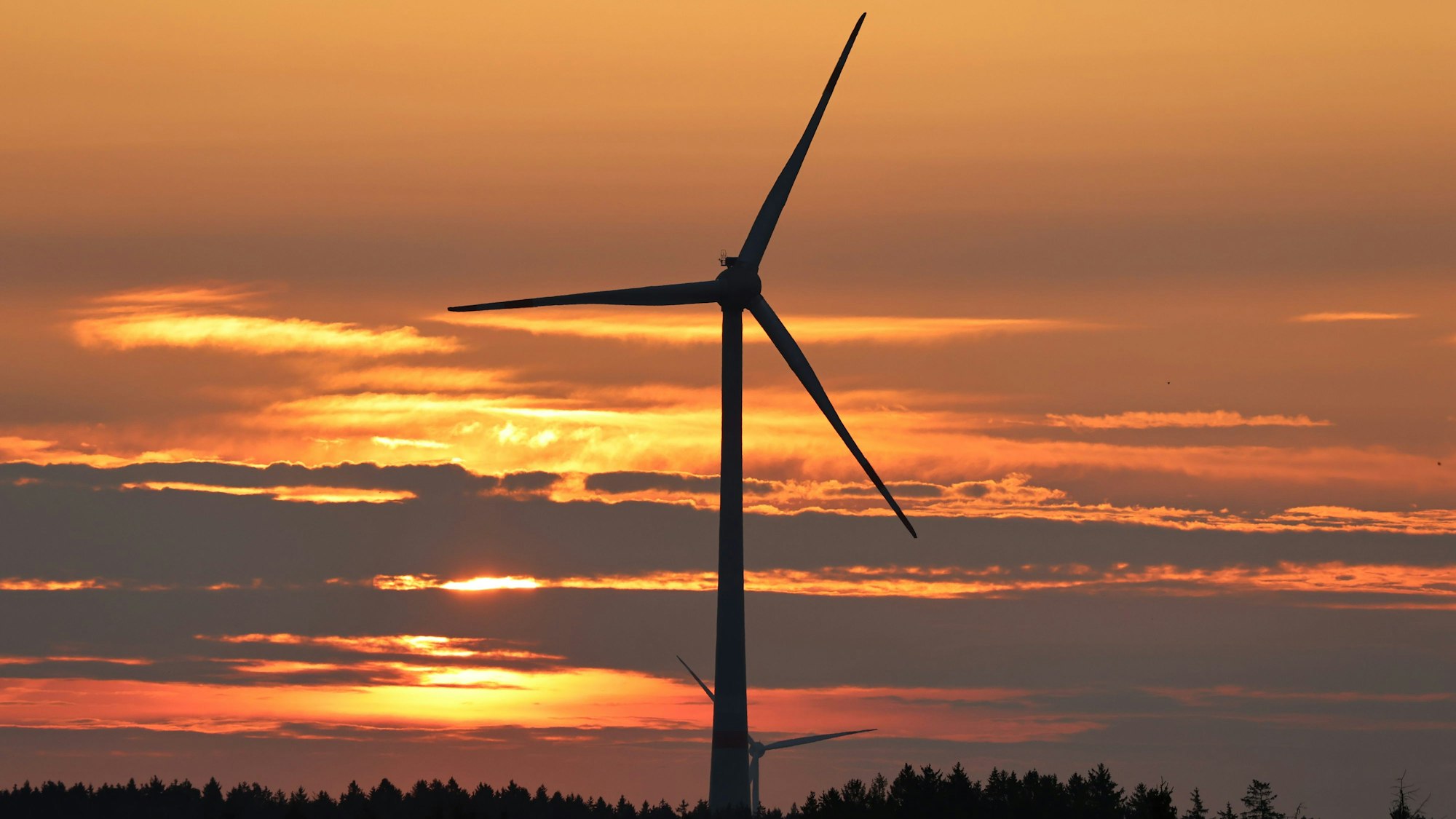 Die Sonne geht an einem bewölkten Himmel hinter einem Windrad unter und taucht den Himmel in orangefarbenes Licht.