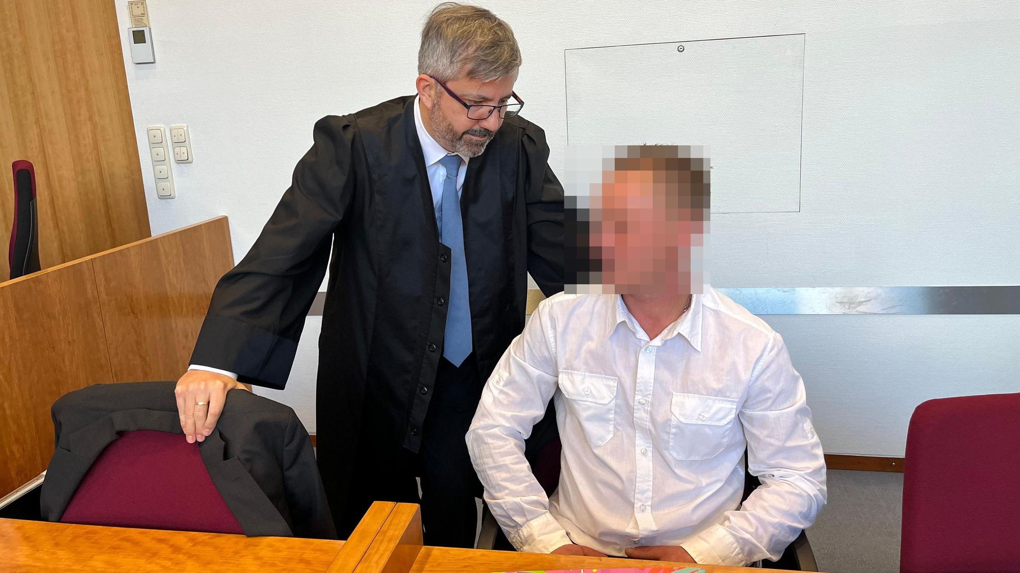 Ein Mann in weißem Oberhemd sitzt auf der Anklagebank, ein Verteidiger steht neben ihm.