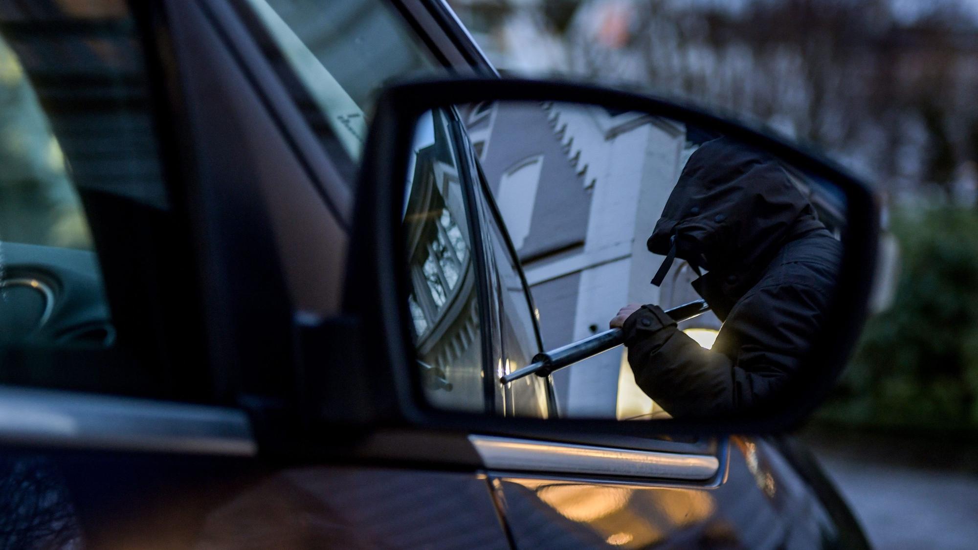 Das Bild zeigt eine vermummte Person, die mit einem Werkzeug versucht, ein Auto aufzubrechen.