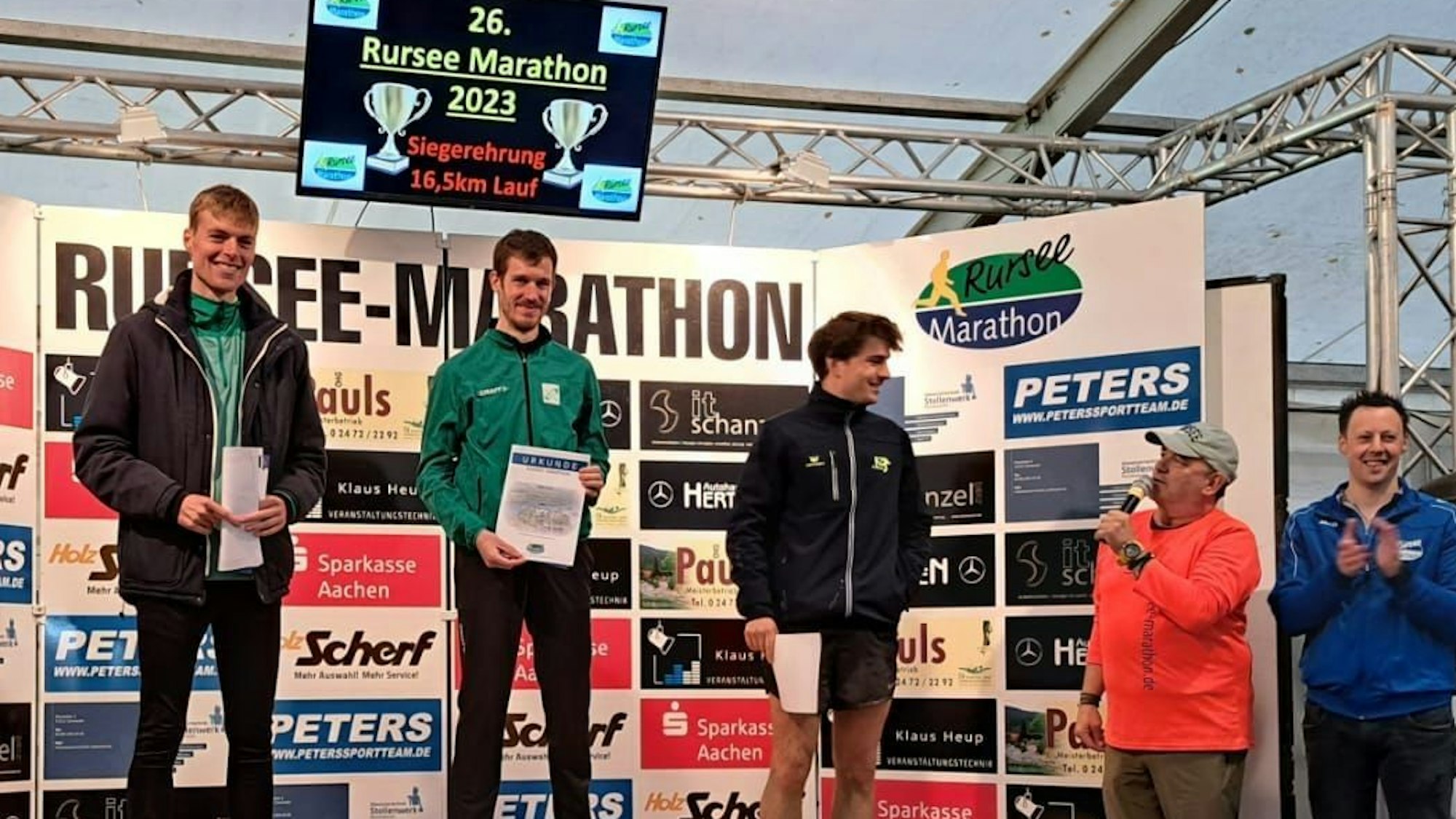 Drei Läufer stehen mit ihren Urkunden auf dem Siegerpodest, ein Moderator stellt sie vor.