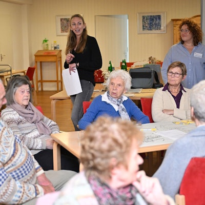 Im Hintergrund stehen die beiden Moderatorinnen neben einer Pinnwand. Im Vordergrund sitzen sieben Teilnehmerinnen und Teilnehmer der Konferenz an einem Tisch.