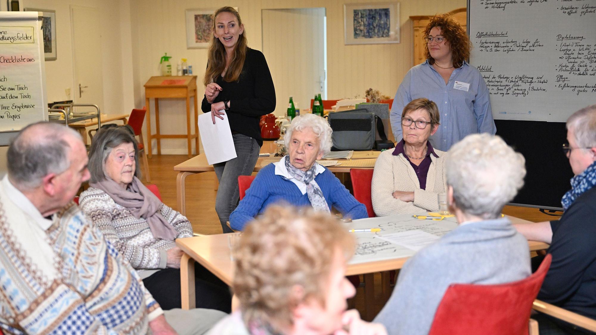 Im Hintergrund stehen die beiden Moderatorinnen neben einer Pinnwand. Im Vordergrund sitzen sieben Teilnehmerinnen und Teilnehmer der Konferenz an einem Tisch.