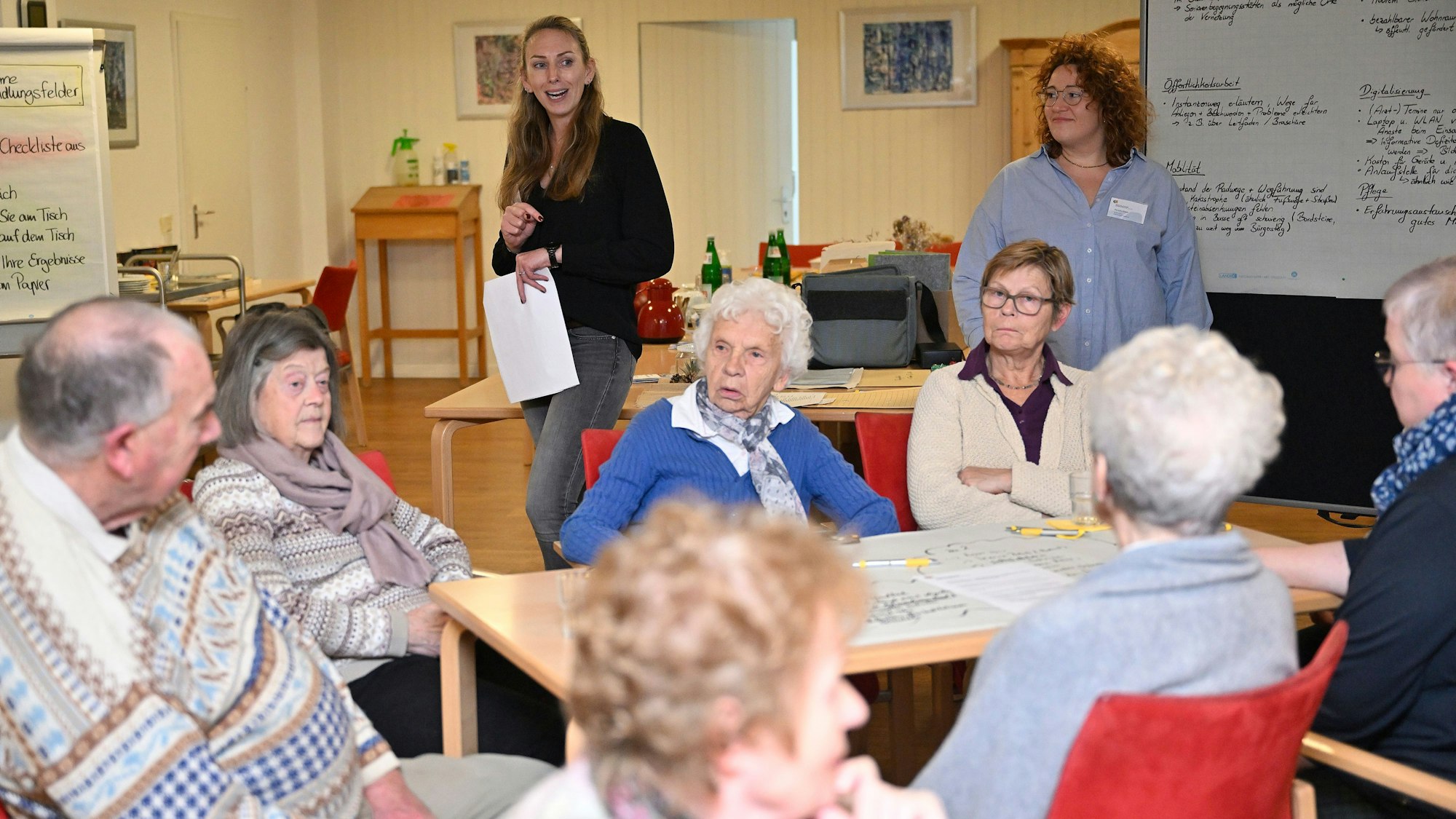 Im Hintergrund stehen die beiden Moderatorinnen neben einer Pinnwand. Im Vordergrund sitzen sieben Teilnehmerinnen und Teilnehmer der Konferenz an einem Tisch.