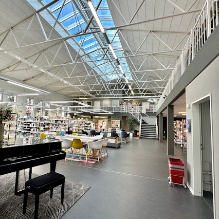 Ein hoher luftiger Raum mit einem Klavier im Vordergrund und Bücherregalen und einer Tischgruppe mit Stühlen ist zu sehen.