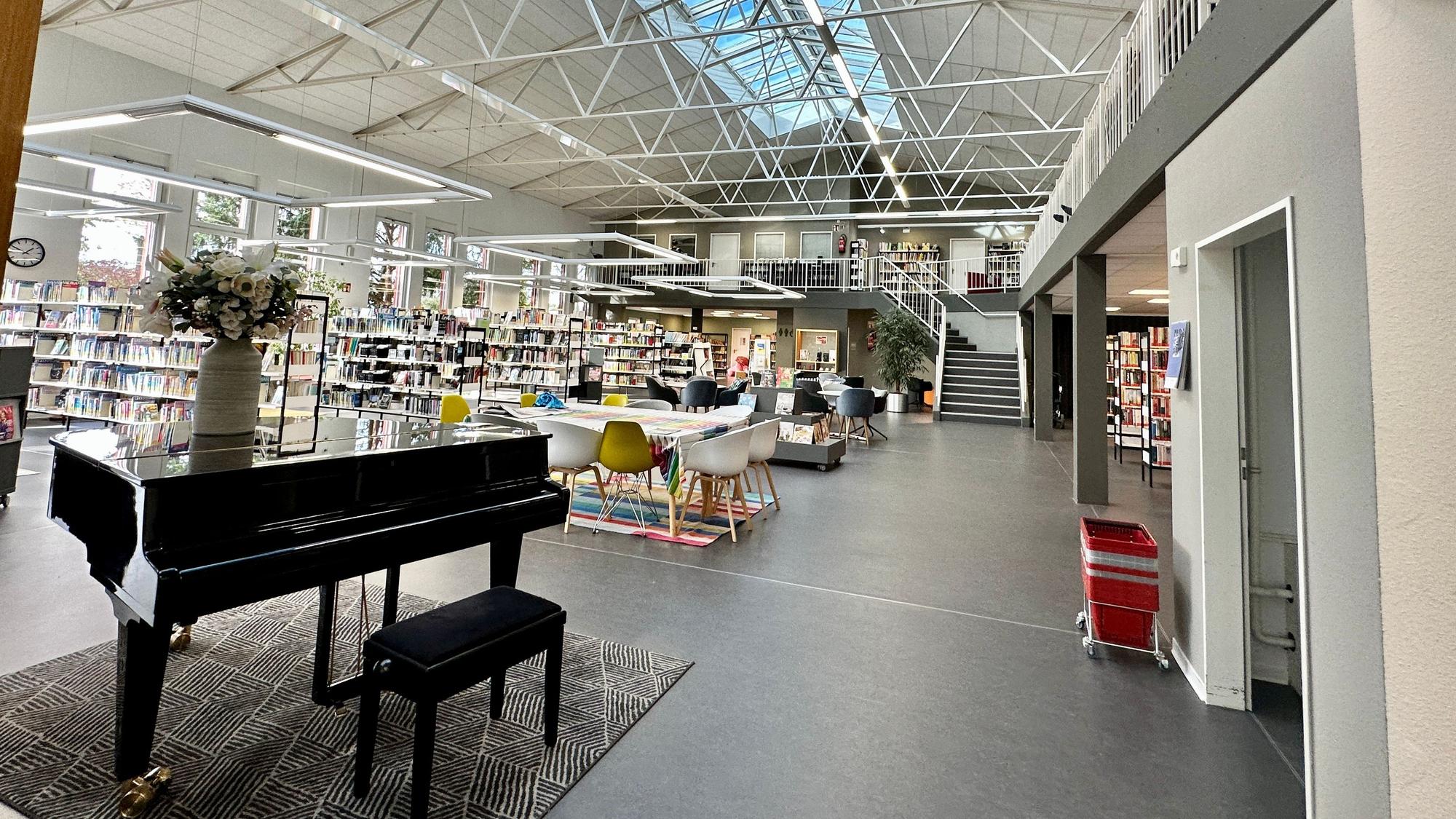 Ein hoher luftiger Raum mit einem Klavier im Vordergrund und Bücherregalen und einer Tischgruppe mit Stühlen ist zu sehen.