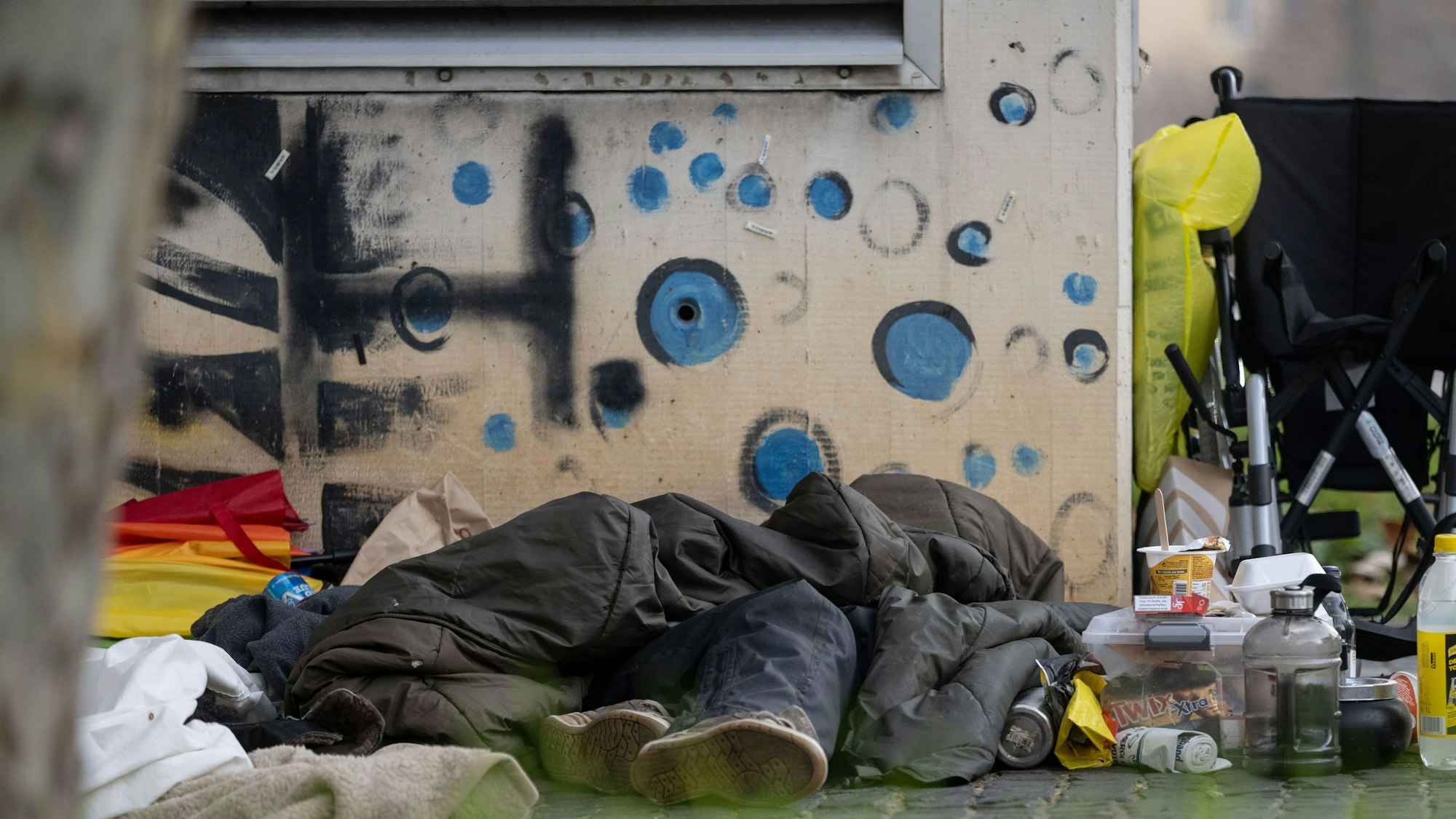 Eine Person liegt an einer Mauer in einem Schlafsack.