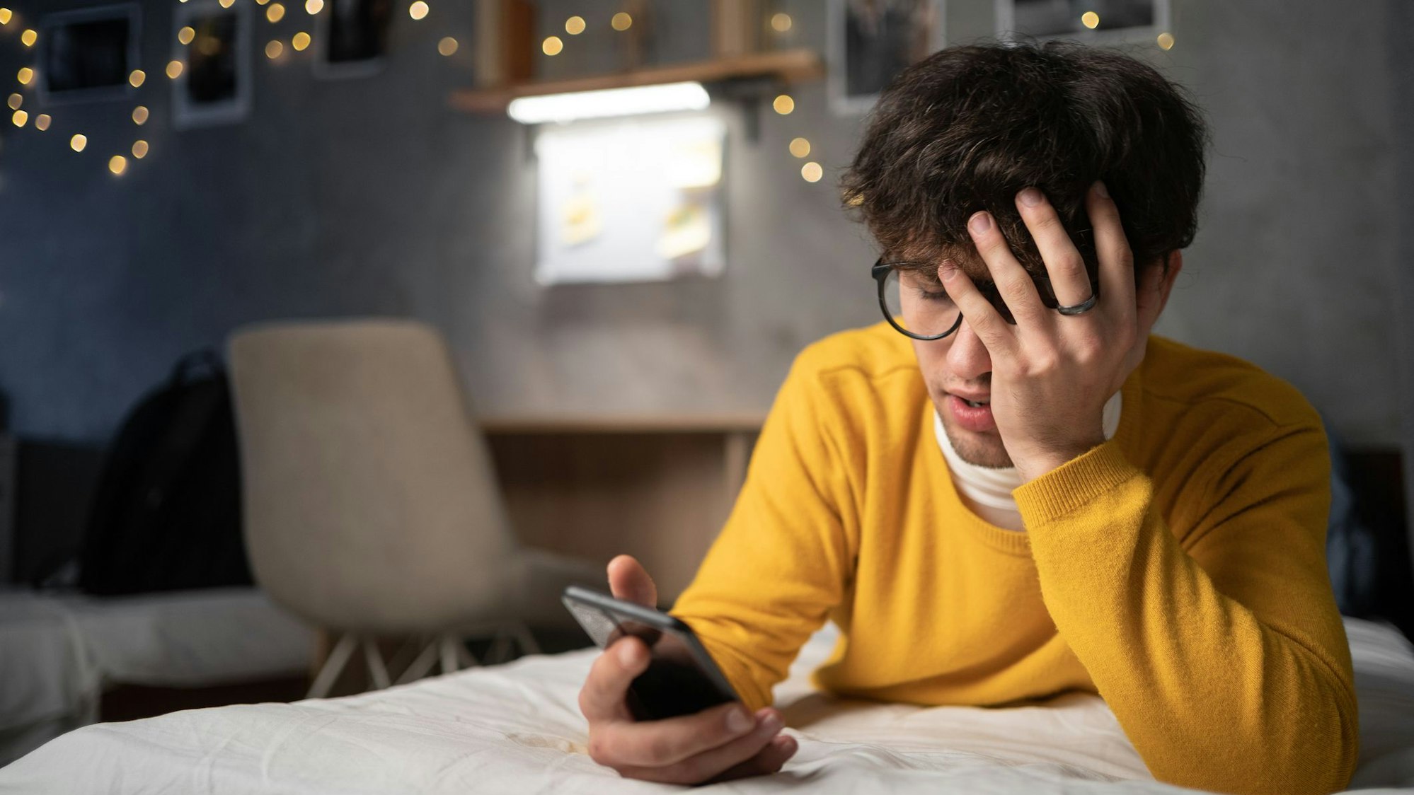 Ein sorgenvoller junger Mann liegt auf dem Bett und schaut auf sein Handy.