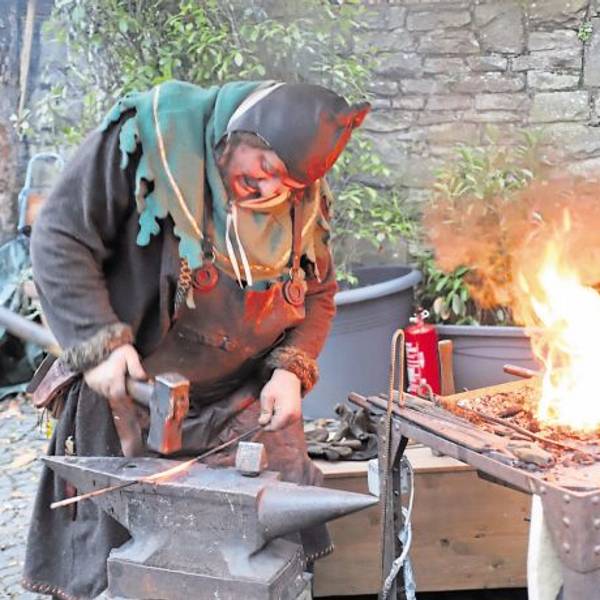 Ein Schmied in mittelalterlichem Kostüm bearbeitet mit einem schweren Hammer ein glühendes Rundeisen auf einem Amboss. Daneben lodert die Esse.