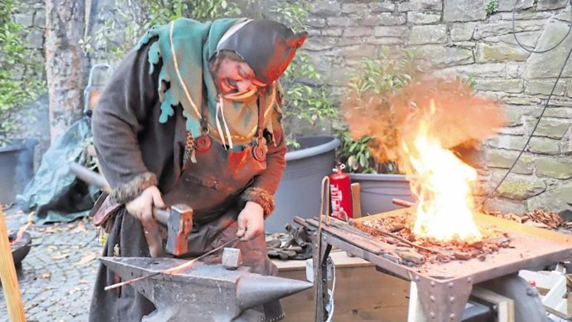 Ein Schmied in mittelalterlichem Kostüm bearbeitet mit einem schweren Hammer ein glühendes Rundeisen auf einem Amboss. Daneben lodert die Esse.