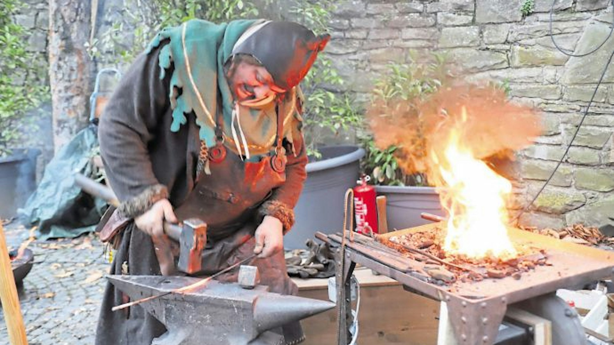 Ein Schmied in mittelalterlichem Kostüm bearbeitet mit einem schweren Hammer ein glühendes Rundeisen auf einem Amboss. Daneben lodert die Esse.