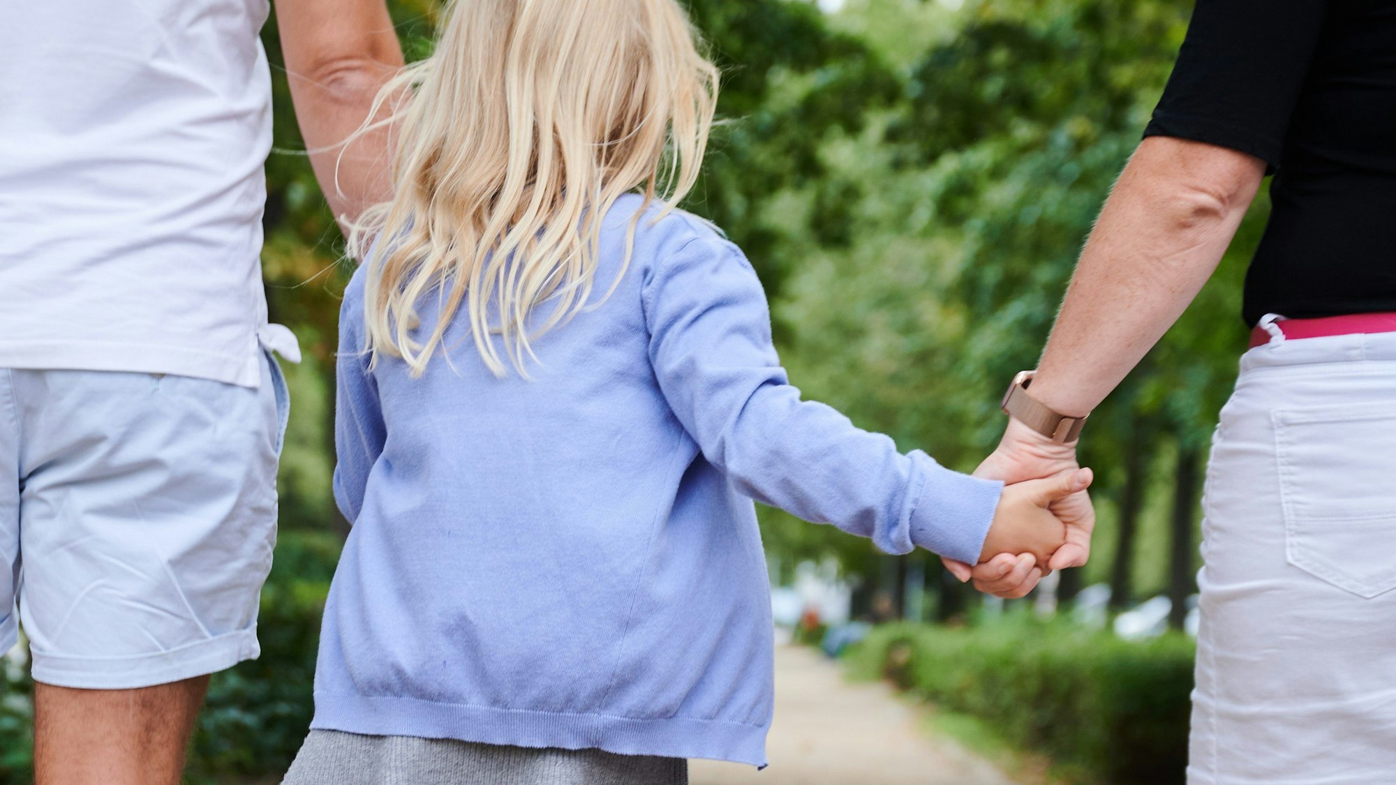 SYMBOLBILD - 27.08.2023, Berlin: Ein Vater und eine Mutter halten die Hände ihrer Tochter, während sie auf einem Gehweg gehen.