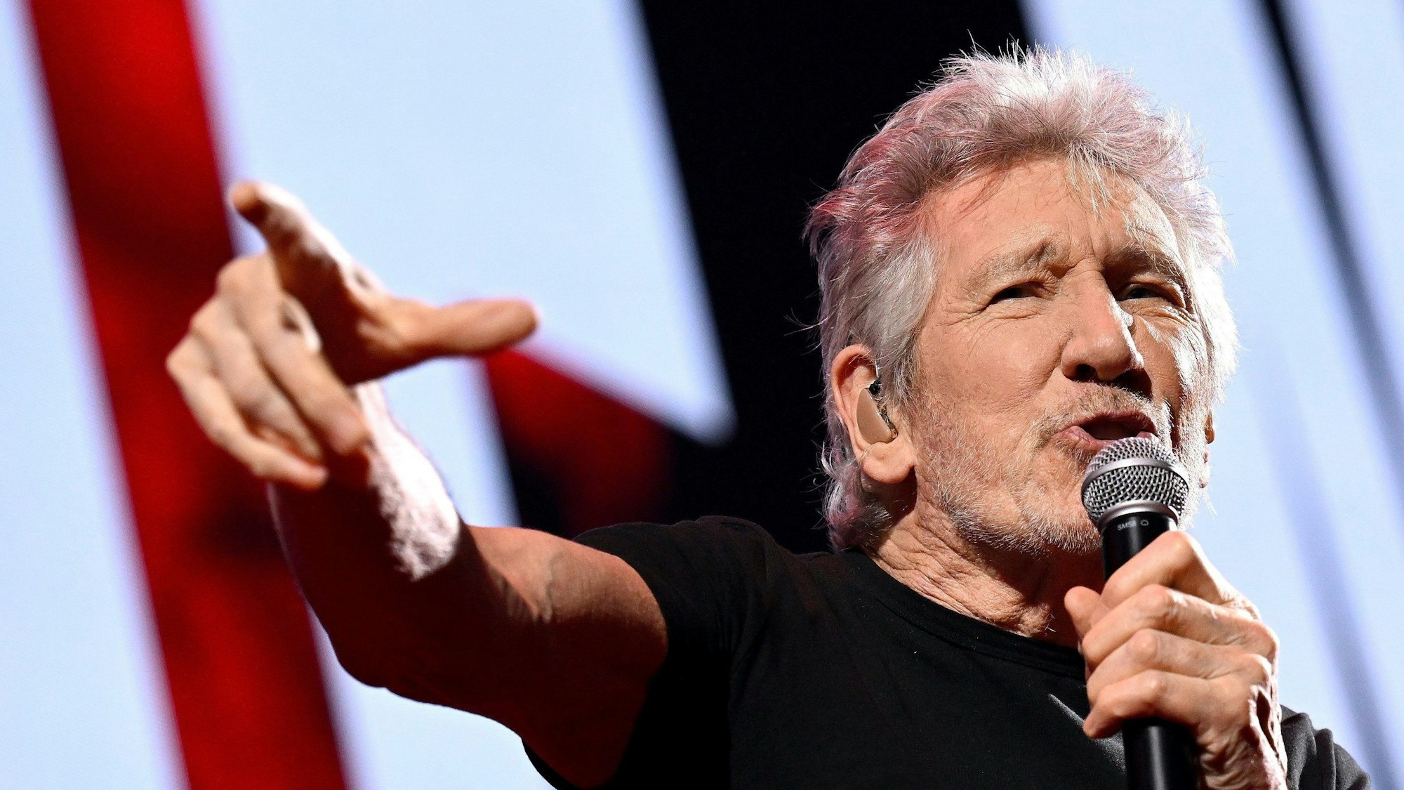 Pink-Floyd-Mitbegründer Roger Waters singt in ein Mikrofon, das er in der Hand hält, während er auf etwas außerhalb des Fotos zeigt.