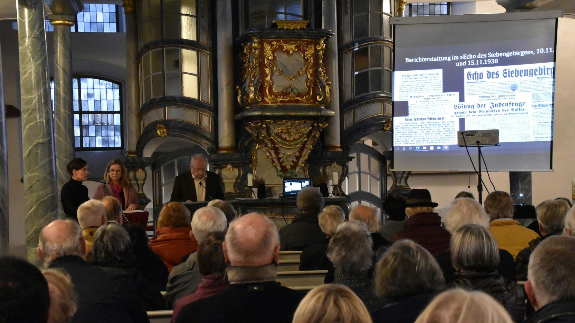 Besucher einer Veranstaltung sitzen in einer Kirche. Menschen sprechen an einem Pult, rechts werden alte Zeitungsseiten auf eine Leinwand projiziert.