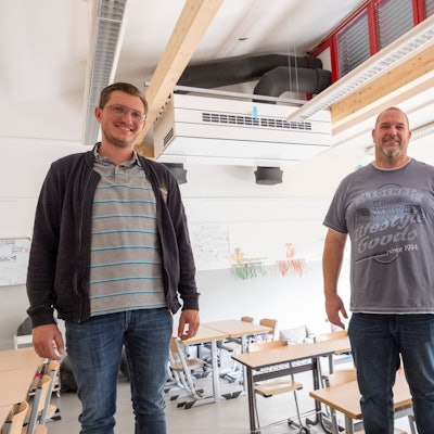 Christoph Breuer (v.l.), Frank Hecker und Andreas Kurth  stehen in einem Klassenraum. Im Hintergrund ist ein Lüftungsgerät an der Decke zu sehen.