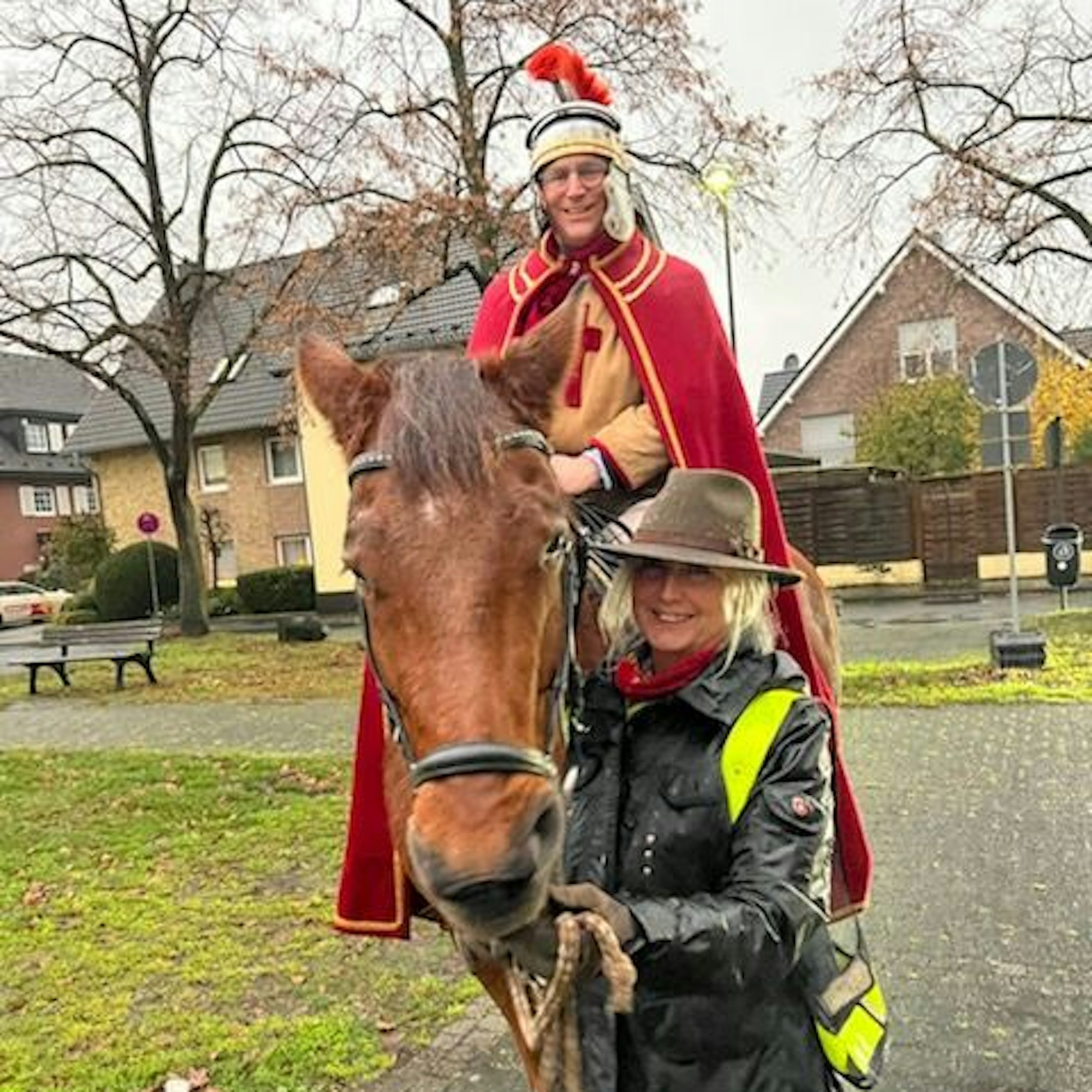 Eine blonde Frau mit Hut und gelber Warnweste führt ein braunes Pferd. Im Sattel sitzt ein Mann in der Kleidung eines römischen Legionärs.