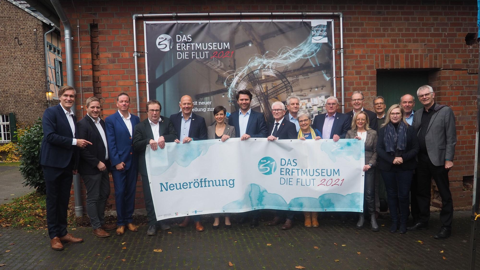 Eine Gruppe steht vor einem Plakat und hält ein Transparent, das auf die Eröffnung der Ausstellung hinweist.