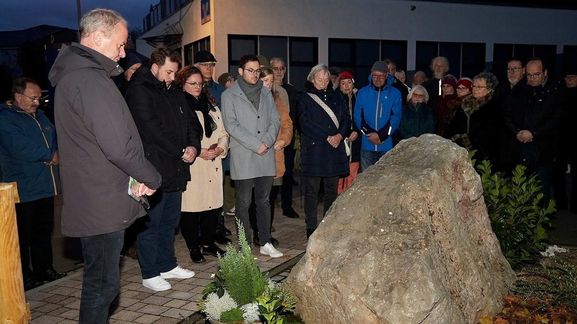 Zahlreiche Menschen stehen um einen mit Blumen geschmückten Gedenkstein herum und halten inne.