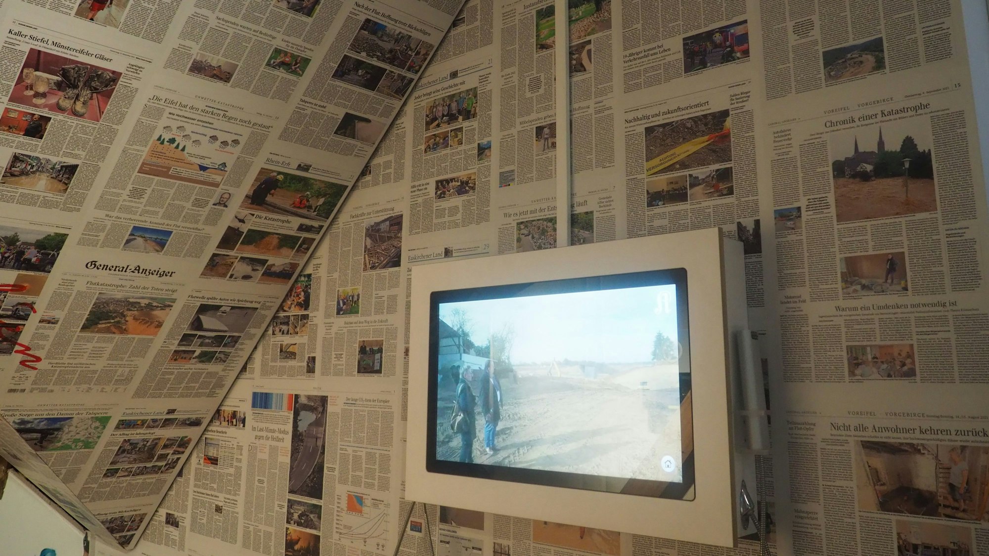 Decke und Wand sind mit Zeitungsseiten bedeckt, davor läuft auf einem Monitor ein Video