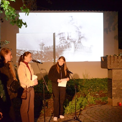 Schülerinnen an Mikrofonen tragen während der Gedenkfeier Texte vor, während ein auf eine Wand projiziertes Bild im Hintergrund die brennende Euskirchener Synagoge zeigt.