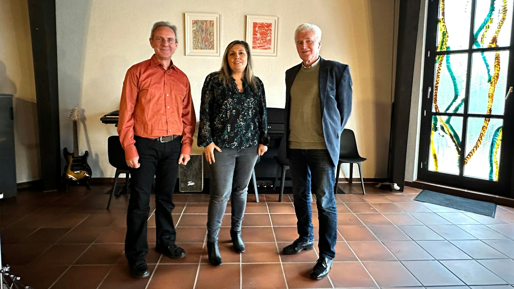 Zwei Männer und eine Frau stehen in einem Raum auf braunen Fliesen, rechts lässt ein buntes Glasfenster Licht herein.