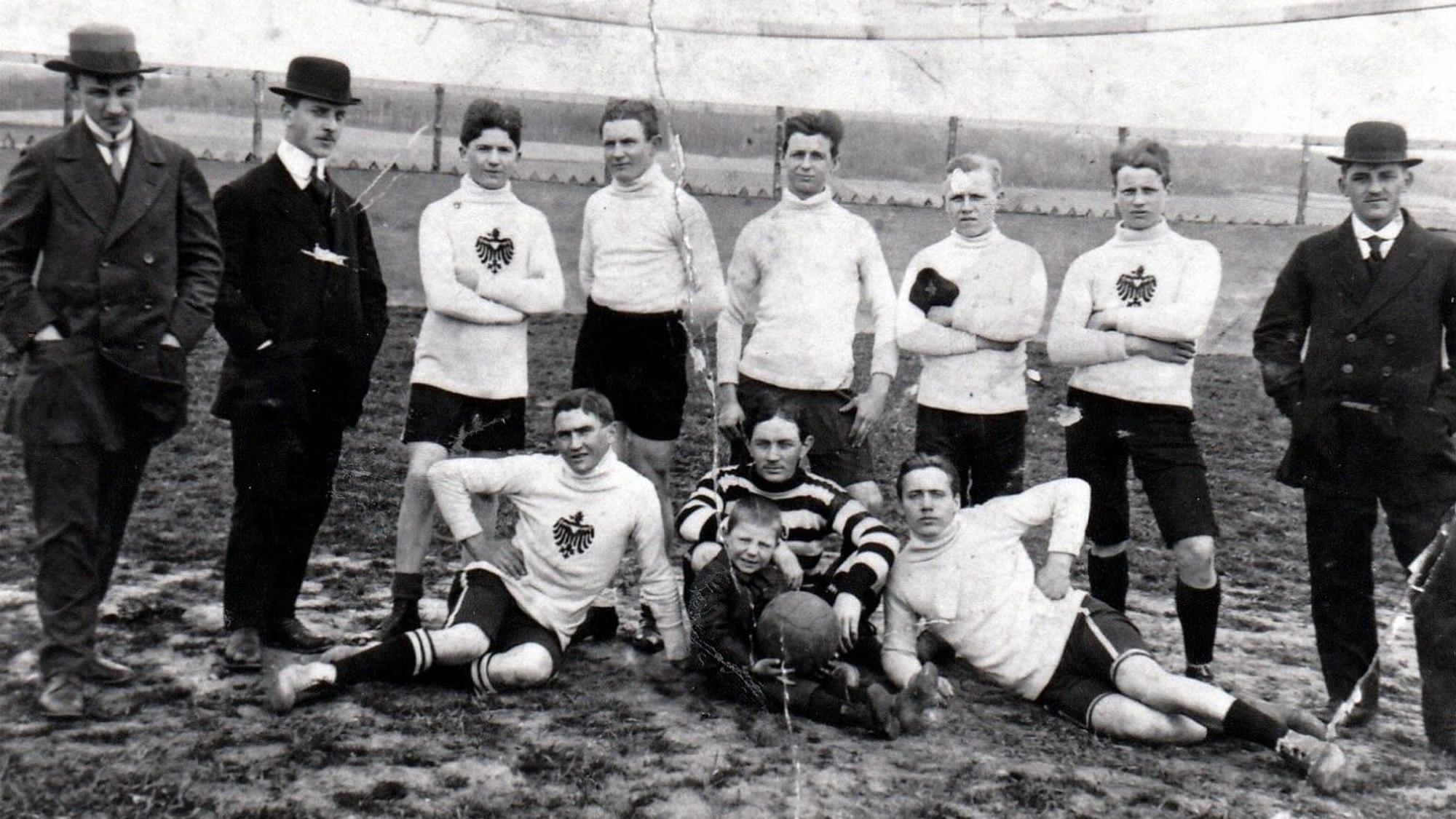 Eine alte Schwarz-Weiß-Fotografie zeigt Fußballer in Trikots und Männer mit Anzug und Melonen-Hüten.