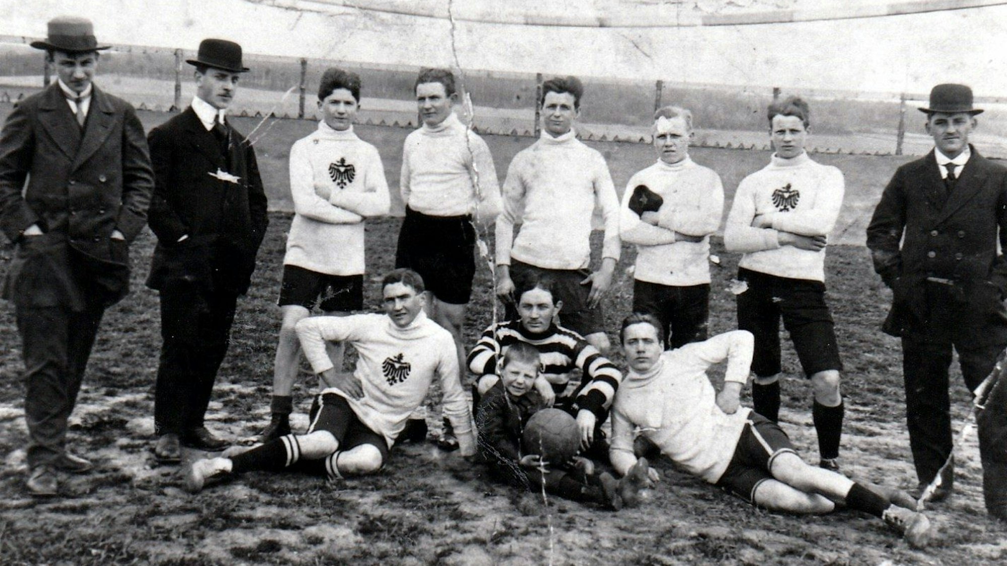 Eine alte Schwarz-Weiß-Fotografie zeigt Fußballer in Trikots und Männer mit Anzug und Melonen-Hüten.