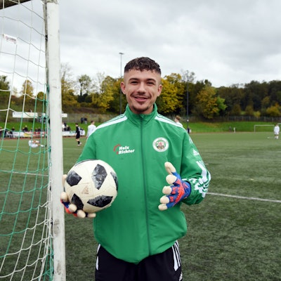 Ein Fußballspieler steht mit Torwarthandschuhen und Ball an einem Tor.