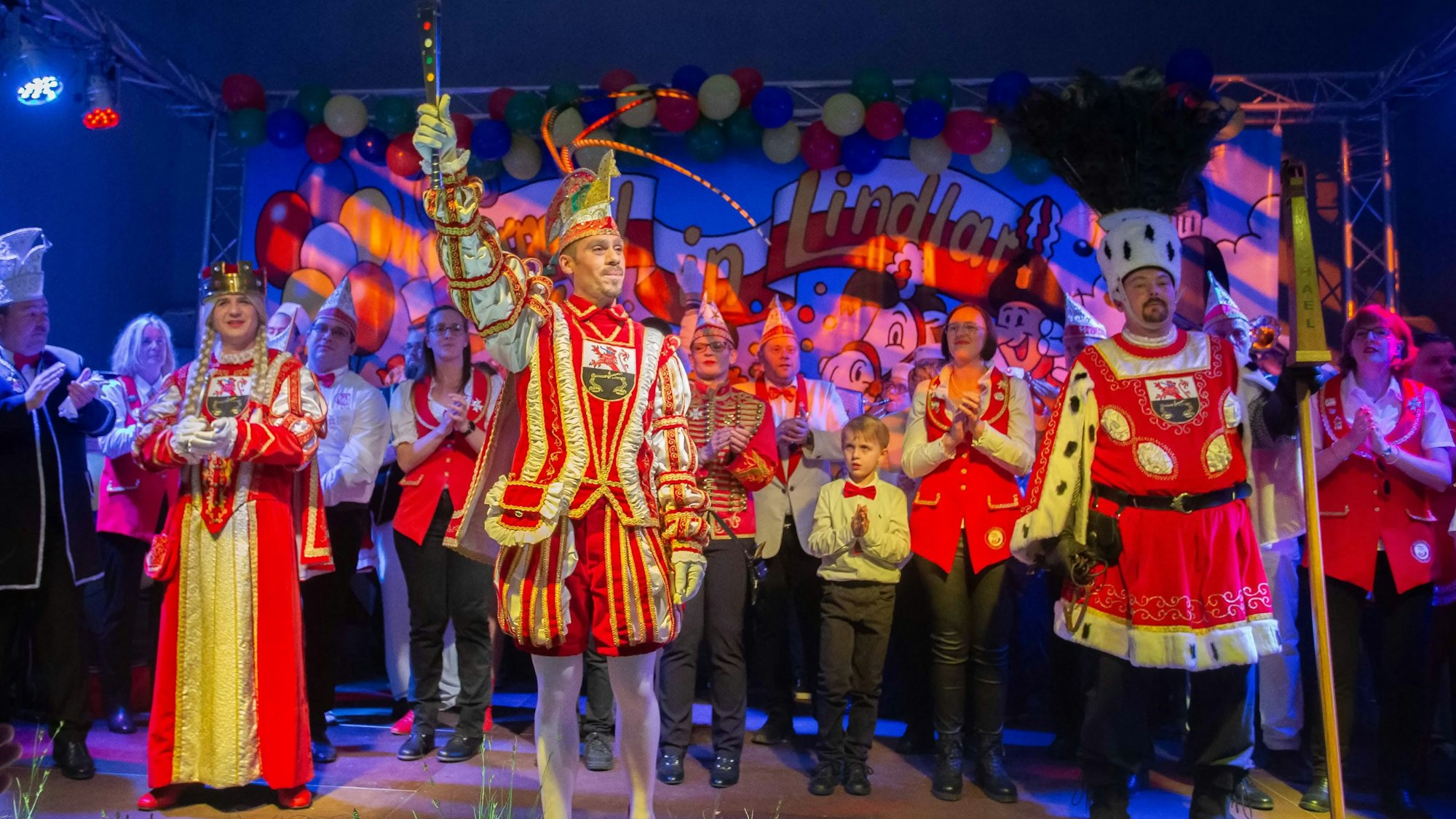 Ein Karnevals-Dreigestirn steht auf einer bunt geschmückten Bühne in einem Festsaal.