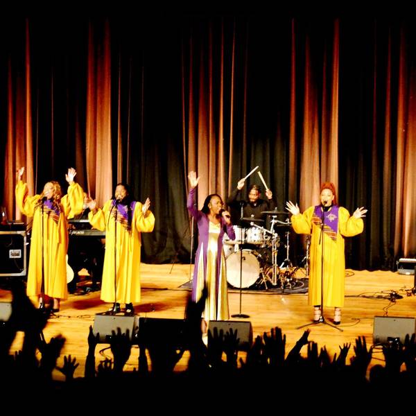 Die Gospelsängerinnen performen in gelben Gewändern auf der Bühnen, während die Zuschauer im Vordergrund die Hände hochrecken.