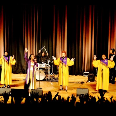 Die Gospelsängerinnen performen in gelben Gewändern auf der Bühnen, während die Zuschauer im Vordergrund die Hände hochrecken.