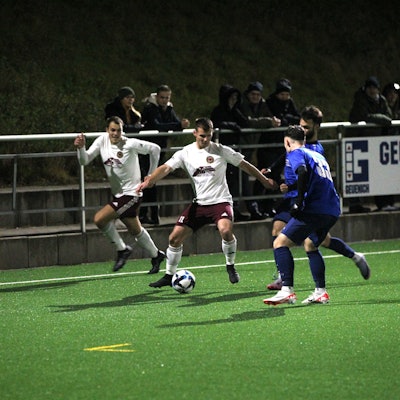 Lommersums Marcel Ebel hat den Ball und wird von zwei Gegenspielern bedrängt. Ein Mitspieler hinterläuft Ebel.