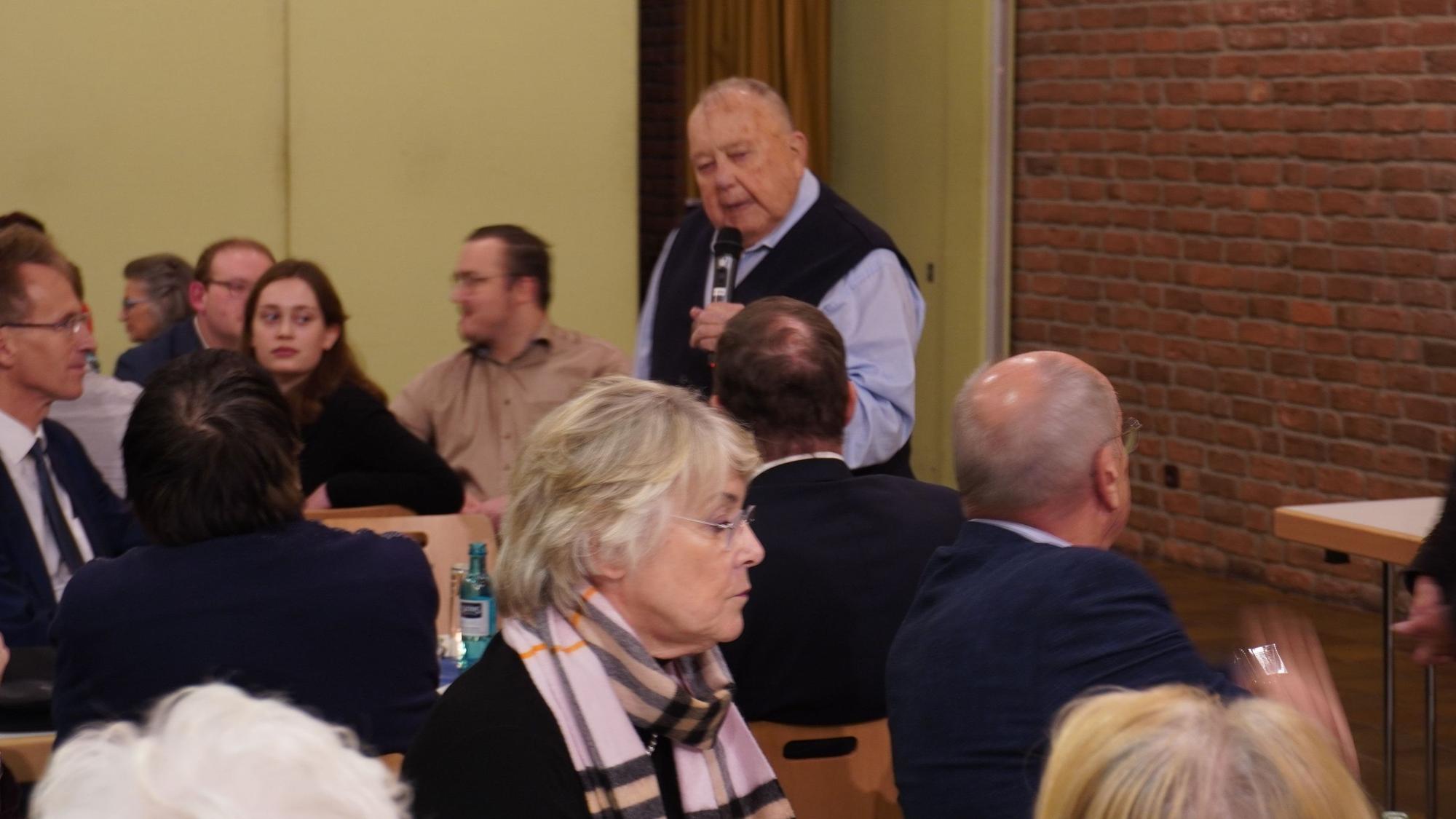 Ein Mann spricht ins Mikrofon, junge und alten Menschen hören zu.