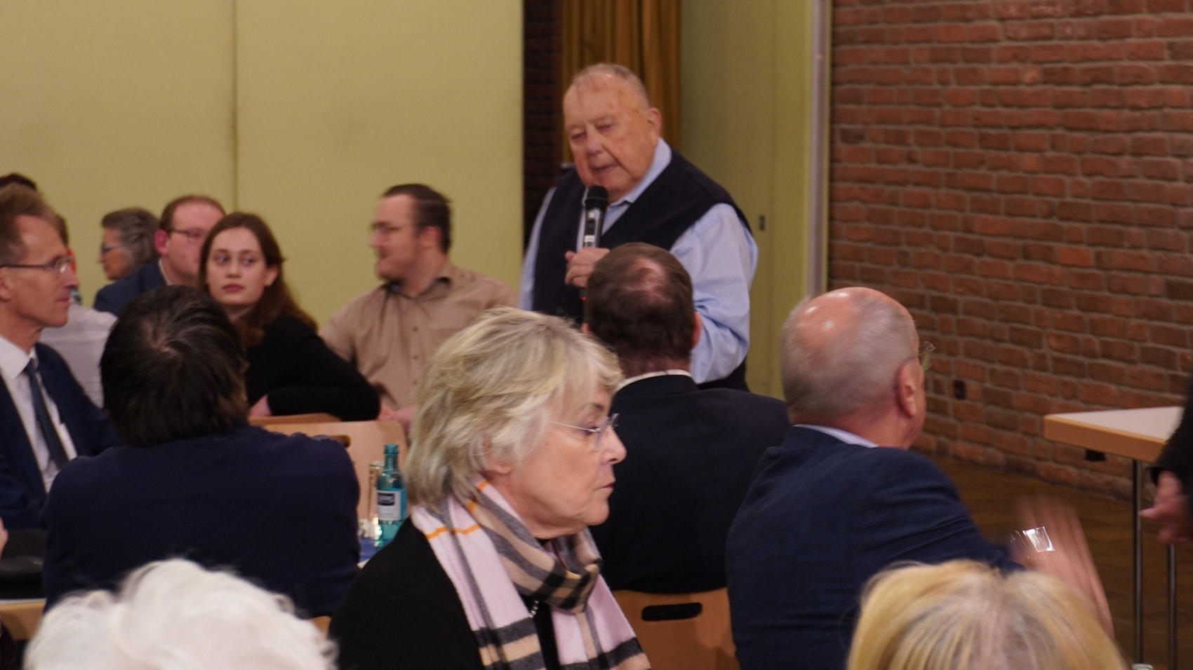 Ein Mann spricht ins Mikrofon, junge und alten Menschen hören zu.