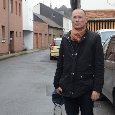 Ein Mann steht auf einer engen Straße. Die Gehwege sind sehr schmal, links und rechts parken Autos.
