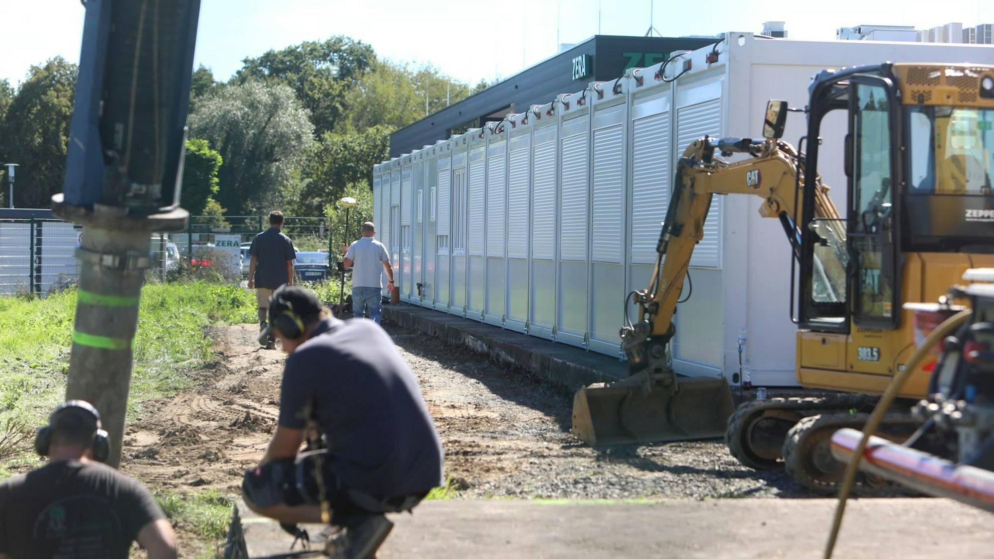 Ein gelber Bagger steht neben einer Reihe von Wohncontainern; davor arbeiten zwei Männer.