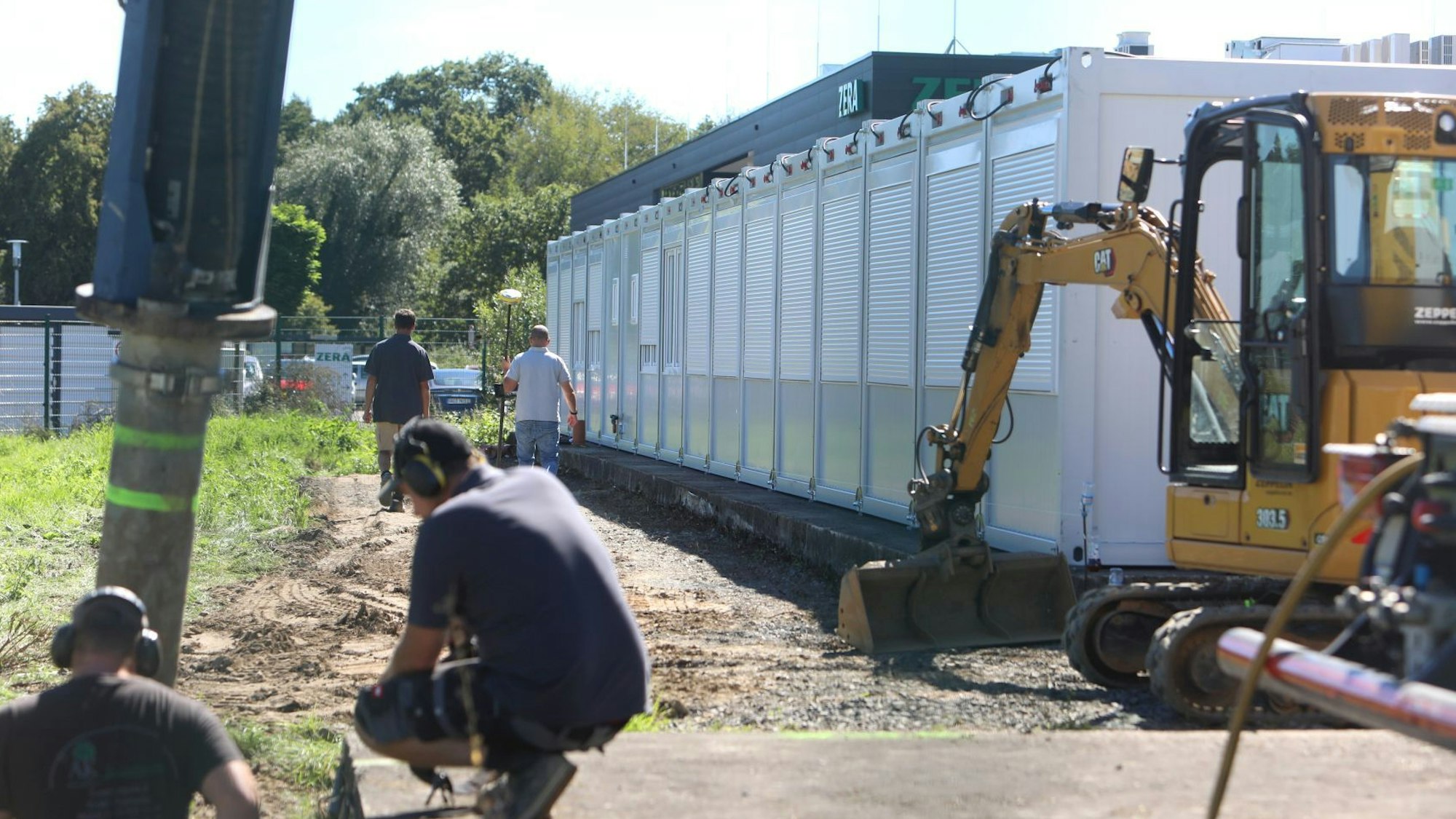 Ein gelber Bagger steht neben einer Reihe von Wohncontainern; davor arbeiten zwei Männer.