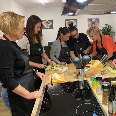 Sechs Frauen mit Schürzen stehen um eine Kochinsel und schnibbeln frische Zutaten wie Paprika und Zwiebeln.