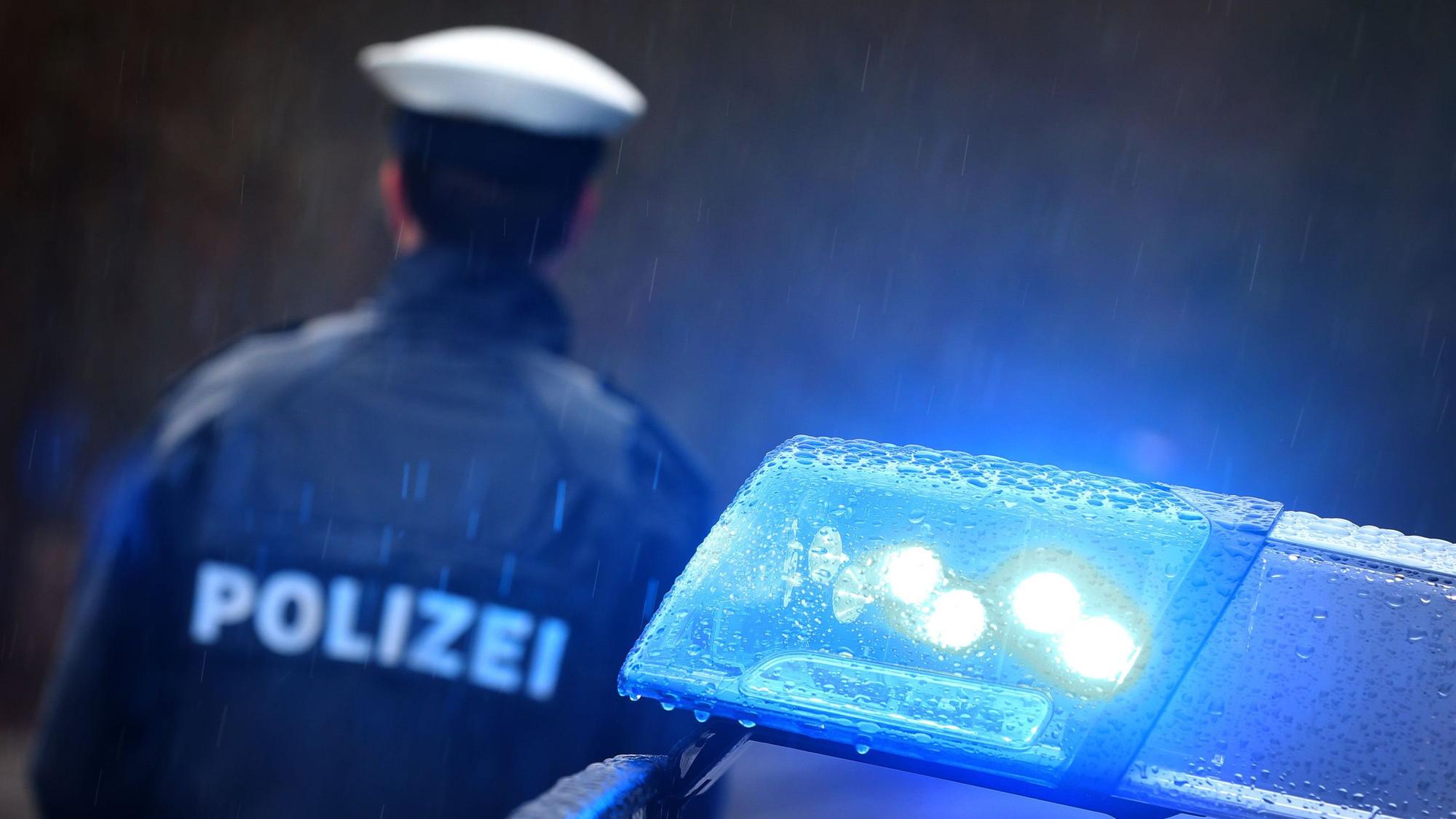 Ein Polizist steht vor einem Streifenwagen dessen Blaulicht aktiviert ist.