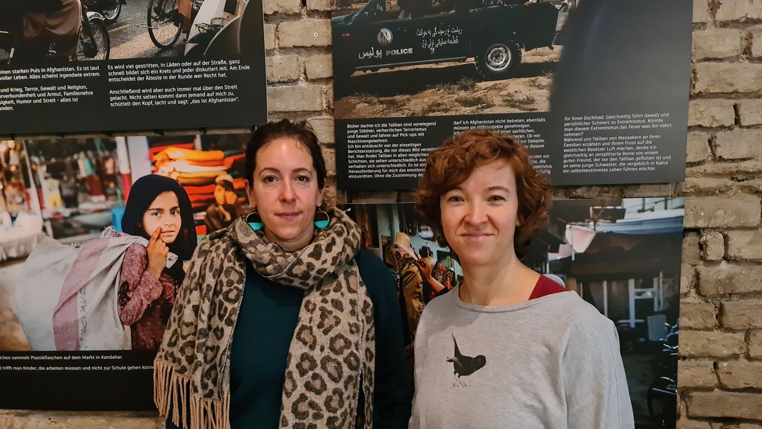 Zwei Frauen stehen nebeneinander vor einer Wand, an der Fotografien von Frauen hängen.