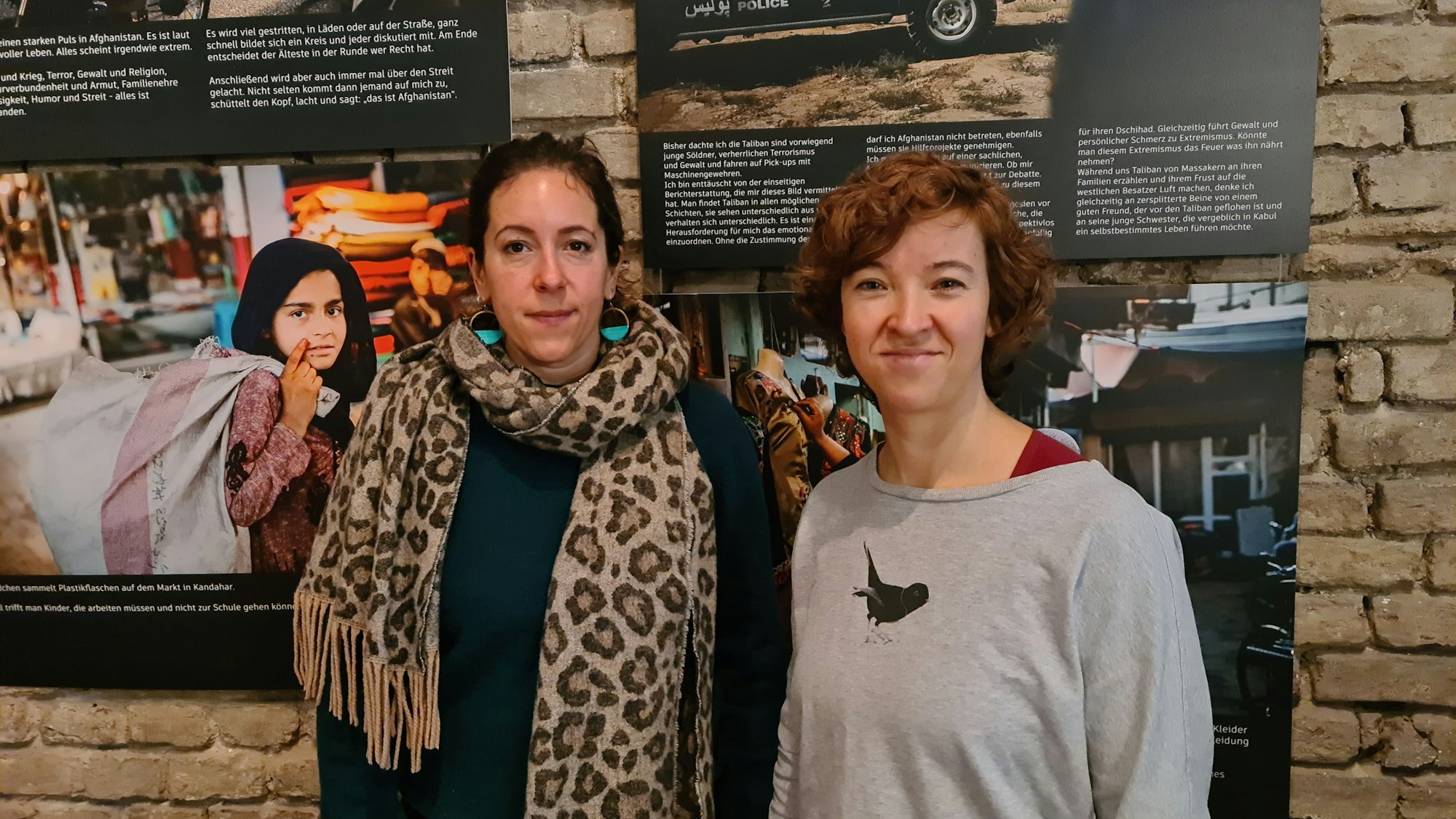 Zwei junge Frauen stehen vor Bildern einer Ausstellung, auf einem ist ein Mädchen zu sehen, das einen schweren Sack trägt.