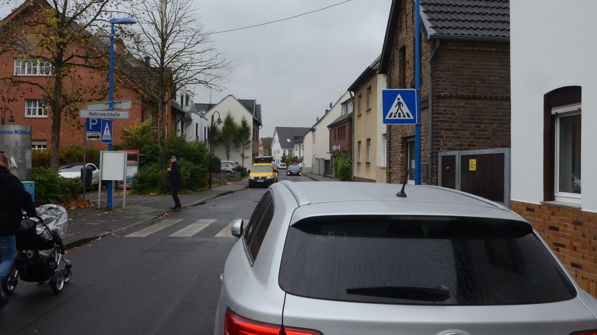 Ein Zebrastreifen im Dorf. Ein Schild weist auf der rechten Straßenseite auf den Übergang hin; ein geparktes Auto verdeckt die Sicht.
