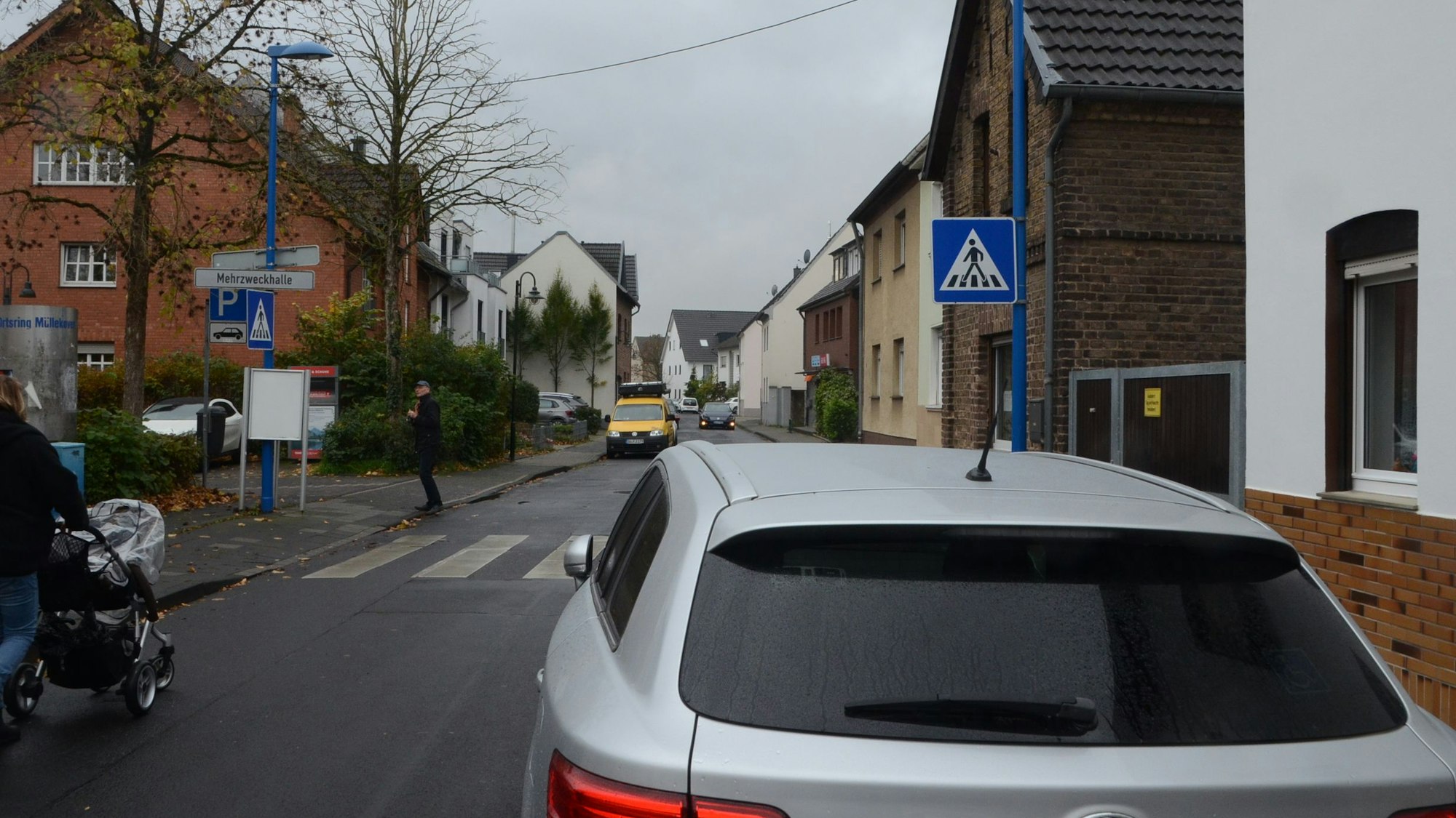 Ein Zebrastreifen im Dorf. Ein Schild weist auf der rechten Straßenseite auf den Übergang hin; ein geparktes Auto verdeckt die Sicht.