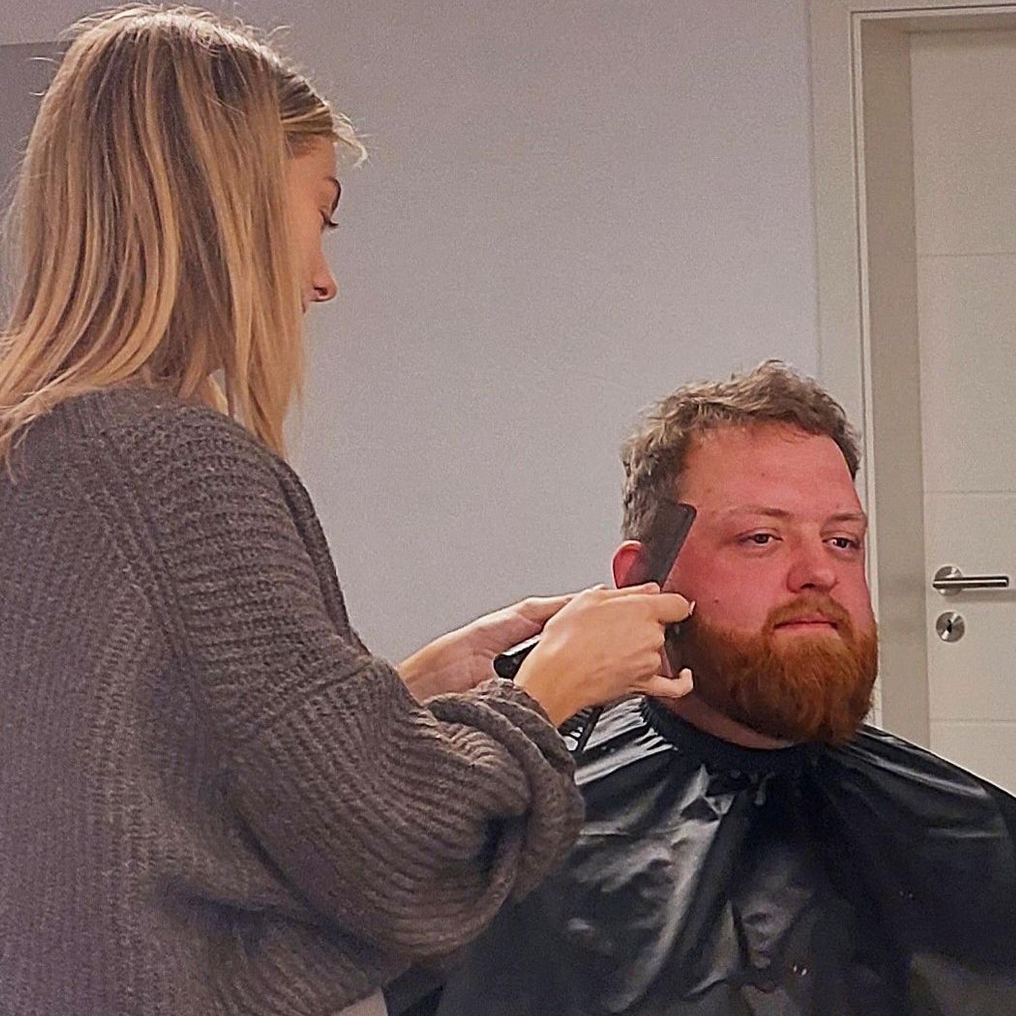 Friseurin Jana Lutterbach nimmt Pierre Hunsicker, der mit einem Umhang im Friseurstuhl sitzt, den Vollbart ab,