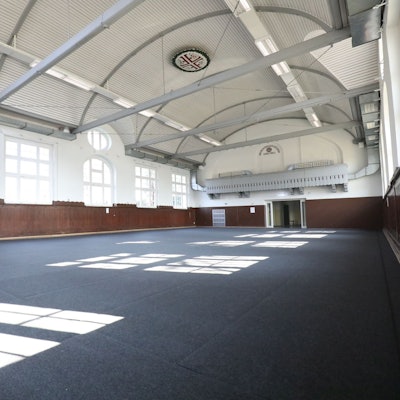 In einer fast leeren Turnhalle mit blauem Boden stehen am linken Rand zwei Personen.