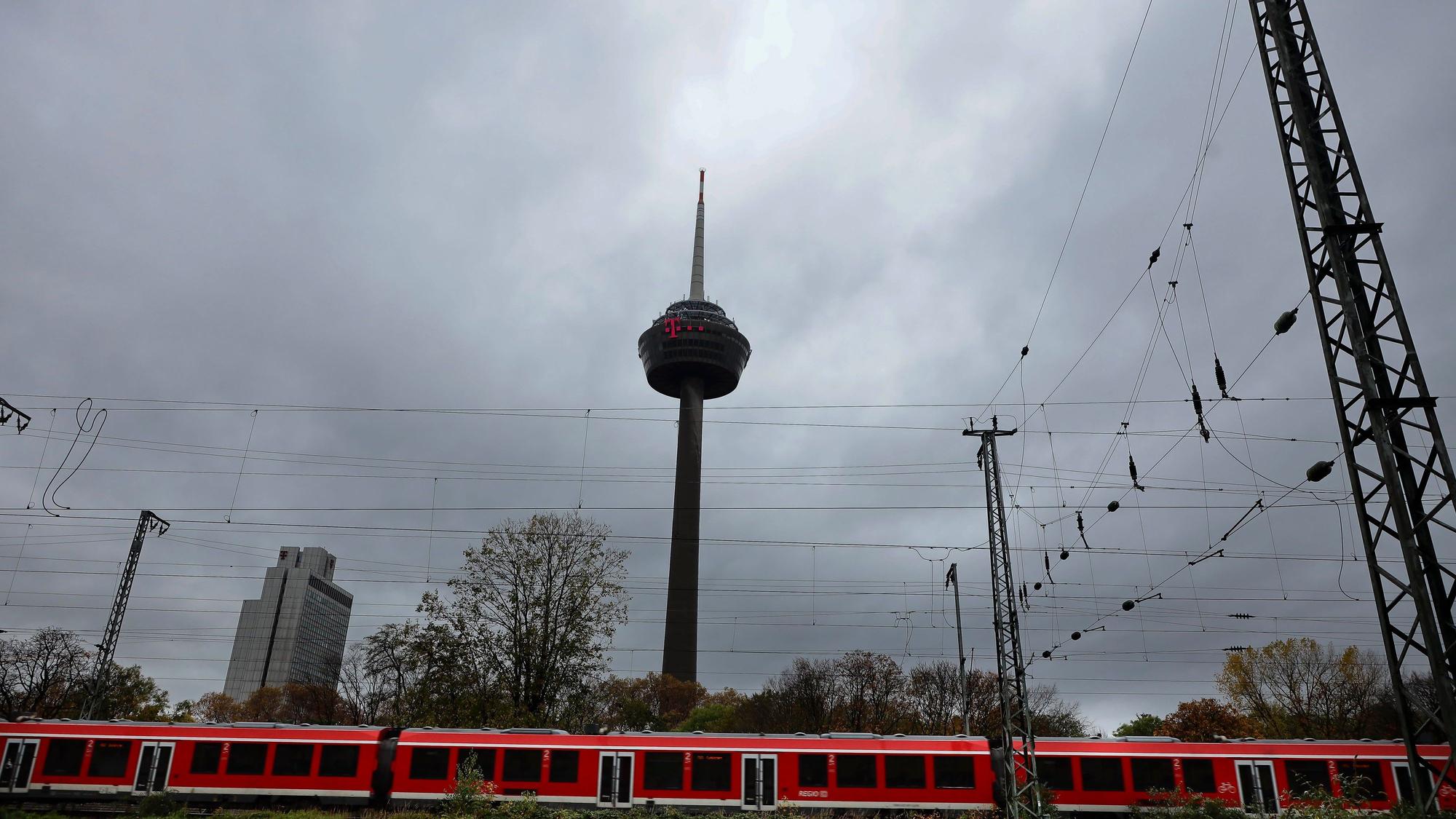 Der Kölner Fernsehturm Colonius ist von dunklen Unwetterwolken umgeben. Im Vordergrund fährt ein Regionalzug auf den Gleisen vorbei. (Archivbild)